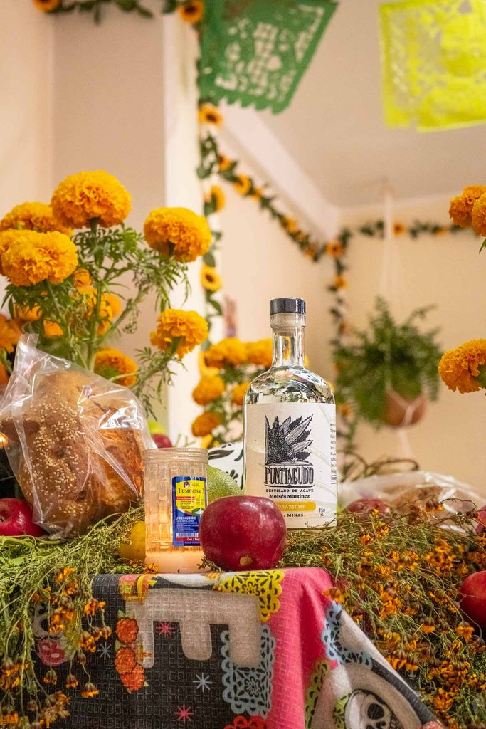 A traditional Day of the Dead offering featuring a bottle of mezcal, marigold flowers, pan de muerto, a red apple, and a lit candle on a decorative cloth.