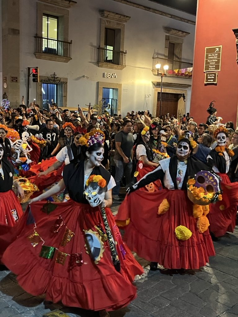 Women in traditional red skirts and catrina makeup dancing in a lively Day of the Dead night parade.